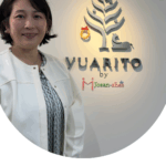 白い壁に「YUARITO by Josan-shes」のロゴが掲げられた日帰り産後ケア施設のエントランス前で、白いジャケットを着た女性（本目さよさん）が微笑んで立っている。ロゴのそばには小さなリースが飾られている。下部には「中央区の日帰り産後ケア施設を見学。赤ちゃんを専門スタッフが丁寧に見守り、手ぶら利用や外出も可能。台東区でも産後ケアの選択肢を広げたい」というコメントが配置されている。